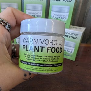 May include: A clear jar of carnivorous plant food with a white lid. The label reads "CARNIVOROUS PLANT FOOD" in large letters. The jar is designed for Venus flytraps, Sundews, and Pitcher plants. Several boxes of the same product are in the background.