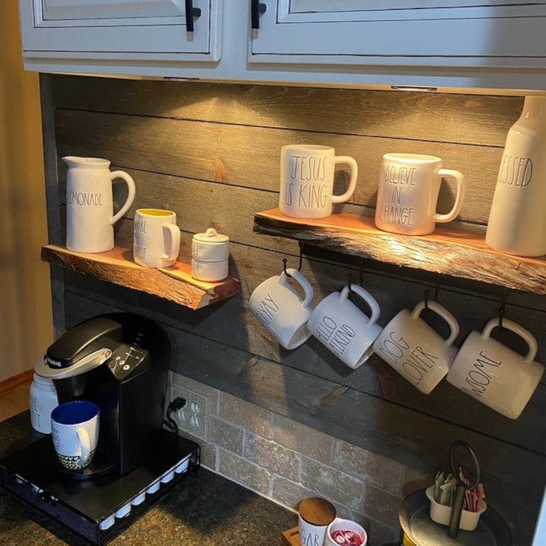 May include: A rustic kitchen with a wooden wall and two shelves. The top shelf has a pitcher labeled "Lemonade", three mugs, and a small jar. The bottom shelf has six mugs hanging from hooks. The mugs have various sayings on them, including "Jesus is King", "Believe in Change", "Hello Friend", "Dog Lover", and "Home".