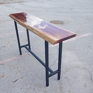 May include: A long, narrow wooden table with a live edge top and a clear epoxy finish. The table has a black metal frame with a simple design. The table is sitting on a gray asphalt surface.