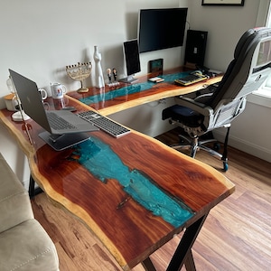 May include: A large, L-shaped desk made of wood with a turquoise blue epoxy river running through the centre. The desk has a black metal frame and is set up in a home office with a laptop, a desktop computer, and a chair.
