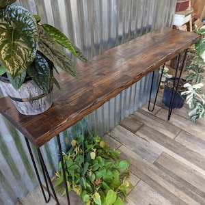 May include: A rustic wooden console table with a dark brown stain. The table has a live edge top and is supported by hairpin legs. The table is in front of a corrugated metal wall and there are plants on the floor.