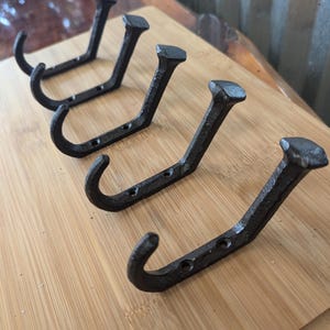 May include: Four black metal hooks with a hammered finish are mounted on a wooden board. The hooks are in a row and are all facing the same direction. The hooks are about 3 inches long and have a slight curve at the top.