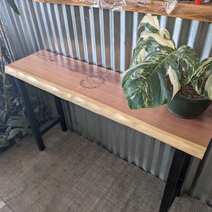 May include: A wooden table with a live edge top and black metal legs. The table has a circular logo burned into the wood that says "El Metro" and "Est. 2017".