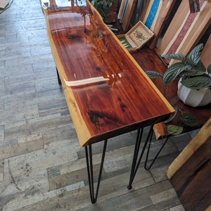May include: A long, rectangular table with a live edge top made of reddish-brown wood. The table has a clear epoxy finish and is supported by four black hairpin legs.