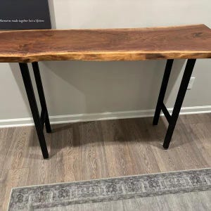 May include: A long, dark brown live edge wood console table with black metal legs. The table is sitting on a wood floor with a rug in front of it.