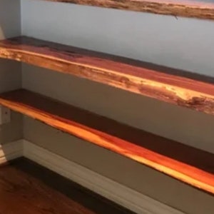 May include: Two wooden shelves with a natural edge, mounted on a wall. The shelves are made of a reddish-brown wood with a natural grain pattern.