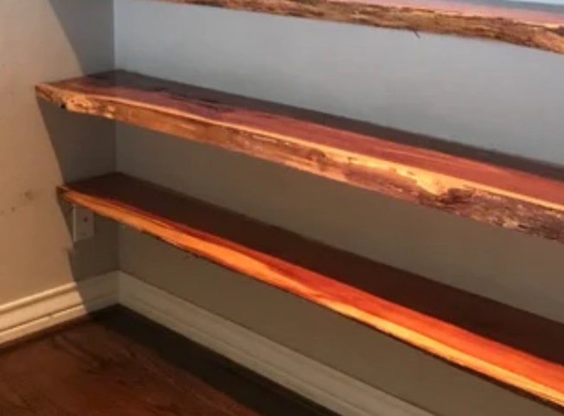 May include: Two wooden shelves with a natural edge, mounted on a wall. The shelves are made of a reddish-brown wood with a natural grain pattern.