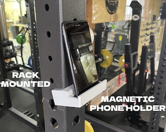 May include: A light gray magnetic phone holder attached to a dark gray metal gym rack. The holder supports a black smartphone in an upright position. The text RACK MOUNTED and MAGNETIC PHONE HOLDER is printed on the image in white capital letters.