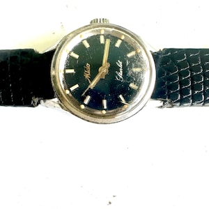 Puede incluir: Reloj plateado vintage con esfera oscura y correa de cuero negro. La esfera del reloj tiene manecillas e indicadores dorados, con las palabras "Mido" y "Starlet". La correa es texturizada.