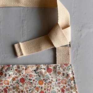 May include: An apron featuring a dense floral pattern with small red, orange, and light blue flowers on a cream background. The apron has wide, beige herringbone-patterned cotton straps that are knotted through a metal ring for adjustment.