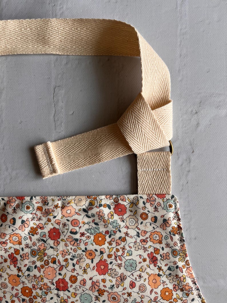 May include: An apron featuring a dense floral pattern with small red, orange, and light blue flowers on a cream background. The apron has wide, beige herringbone-patterned cotton straps that are knotted through a metal ring for adjustment.