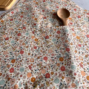 May include: A kitchen apron featuring a dense floral pattern in shades of coral, orange, and light blue on a cream background. The apron includes a large front pocket containing a wooden spoon and has light-colored fabric neck straps.