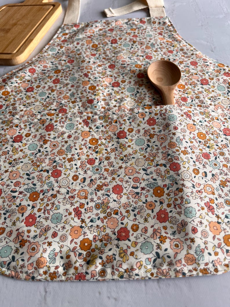 May include: A kitchen apron featuring a dense floral pattern in shades of coral, orange, and light blue on a cream background. The apron includes a large front pocket containing a wooden spoon and has light-colored fabric neck straps.