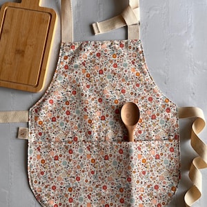 May include: A kitchen apron featuring a cream-colored fabric with a dense floral pattern in shades of orange, red, and blue. The apron includes a large front pocket, beige cotton neck and waist ties, and a wooden spoon tucked into the pocket.