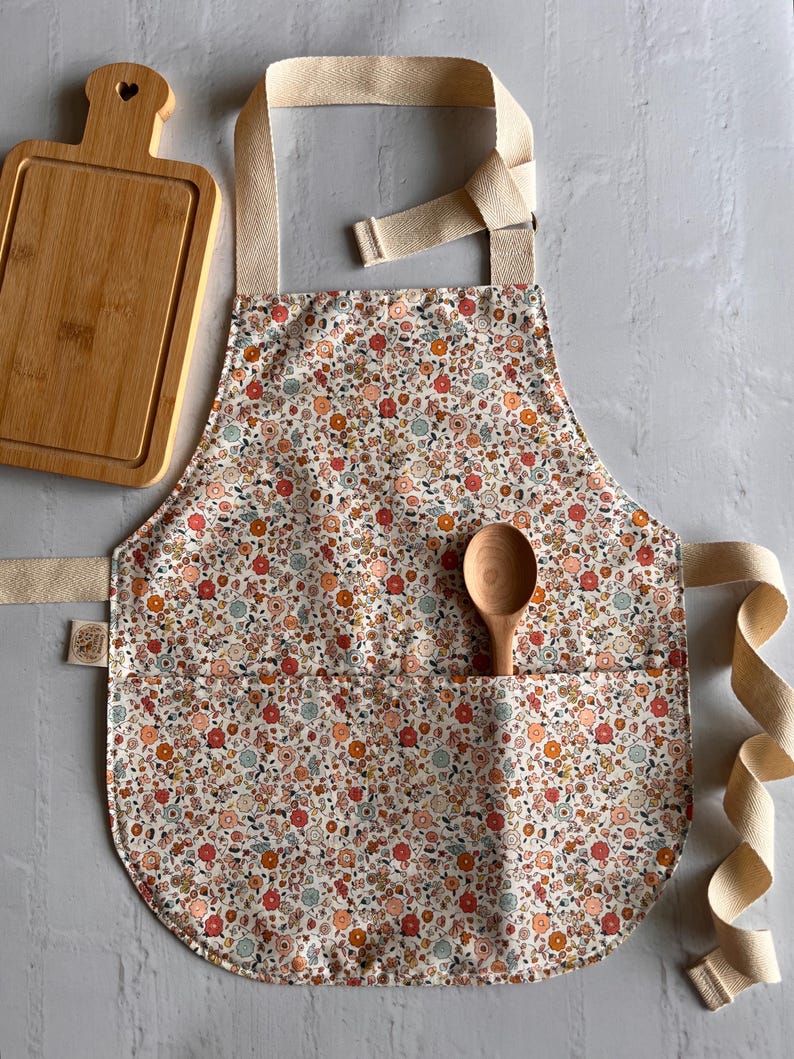 May include: A kitchen apron featuring a cream-colored fabric with a dense floral pattern in shades of orange, red, and blue. The apron includes a large front pocket, beige cotton neck and waist ties, and a wooden spoon tucked into the pocket.