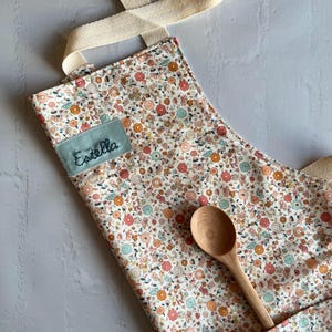 May include: A white apron with a floral print in shades of pink, orange, and green. The apron has a large pocket on the front and a name tag that says "Estella". A wooden spoon is lying on top of the apron.