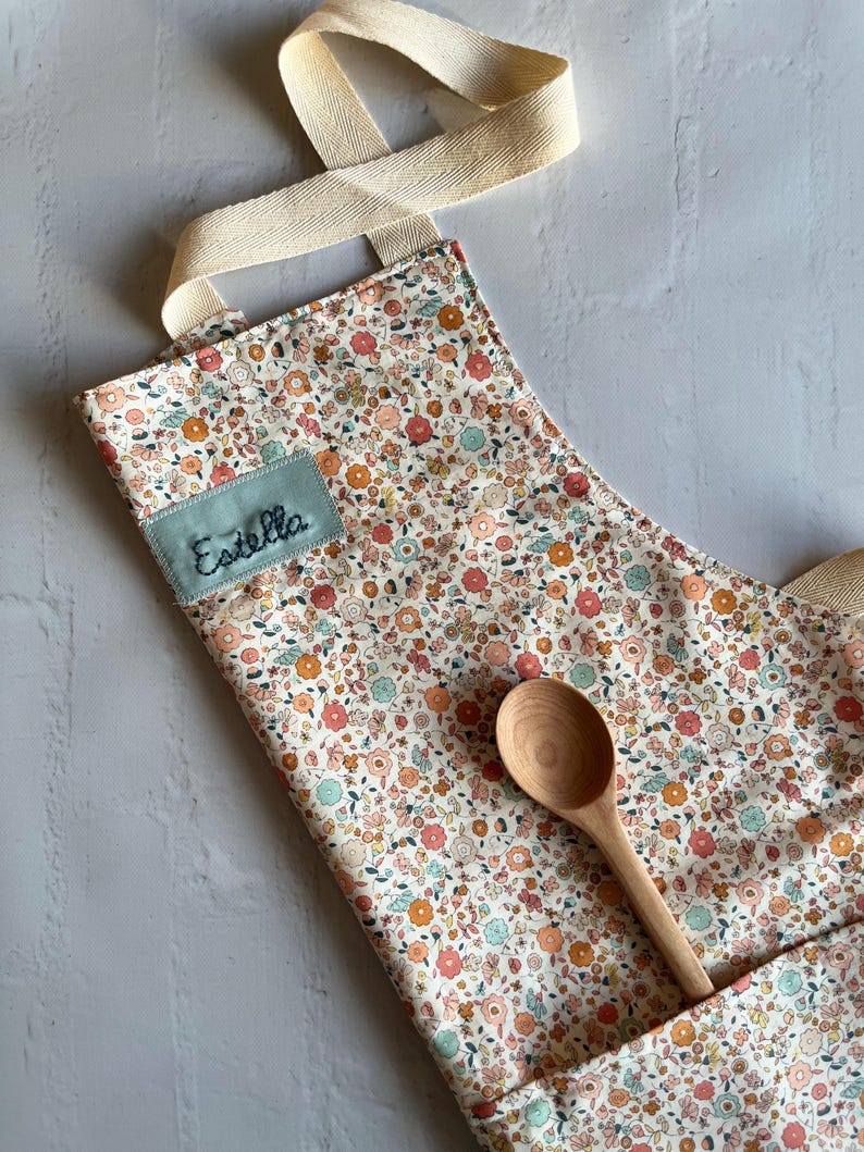 May include: A white apron with a floral print in shades of pink, orange, and green. The apron has a large pocket on the front and a name tag that says "Estella". A wooden spoon is lying on top of the apron.
