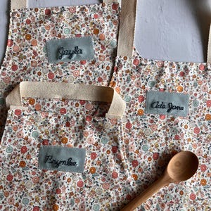 May include: Three floral aprons with beige straps and embroidered name patches. The aprons feature a colorful flower pattern on a white background. A wooden spoon rests on one of the aprons.