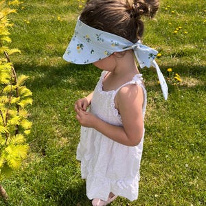 May include: A light blue visor with a floral pattern is tied in a bow. The visor is worn by a person wearing a white eyelet dress and pink shoes. The background is a green grassy field with a white fence.