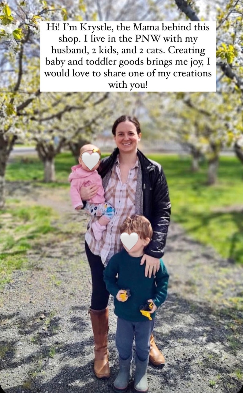 May include: A woman with two children in a park setting. The woman is wearing a plaid shirt, a black jacket, and brown boots. The children are wearing a pink sweater, blue jeans, and green boots. The woman is holding the younger child in her arms. The older child is standing next to her. The background is a blurred image of trees and grass.