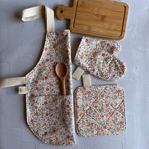 May include: A kitchen set featuring a floral print apron with a front pocket, a matching oven mitt, a square quilted pot holder, a wooden spoon, and a rectangular bamboo cutting board with a heart-shaped cutout on the handle.