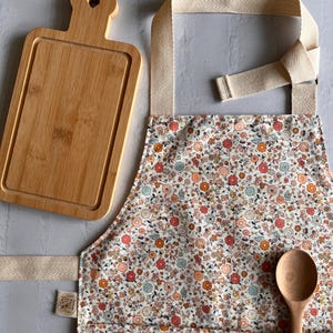 May include: A rectangular bamboo cutting board with a heart-shaped cutout in the handle, a floral print apron with beige herringbone straps, and a wooden spoon. The apron features a small fabric tag on the side and a front pocket holding the spoon.