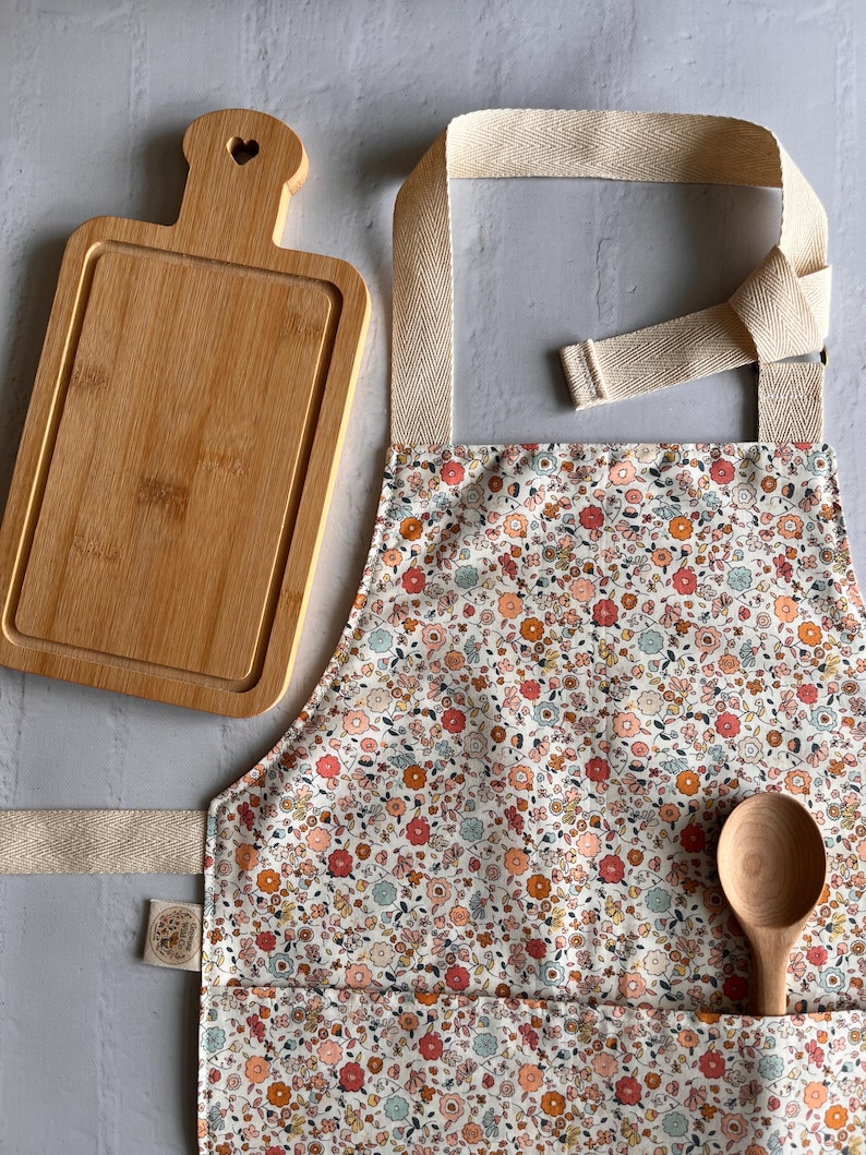 May include: A rectangular bamboo cutting board with a heart-shaped cutout in the handle, a floral print apron with beige herringbone straps, and a wooden spoon. The apron features a small fabric tag on the side and a front pocket holding the spoon.
