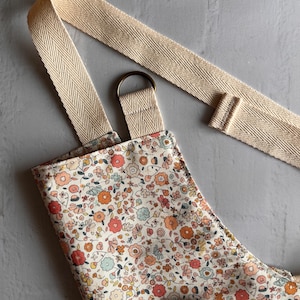 May include: An apron featuring a dense floral pattern with small red, orange, pink, and light blue flowers on a cream background. It includes wide, beige herringbone-patterned cotton straps and a bronze-colored metal D-ring for adjustment.