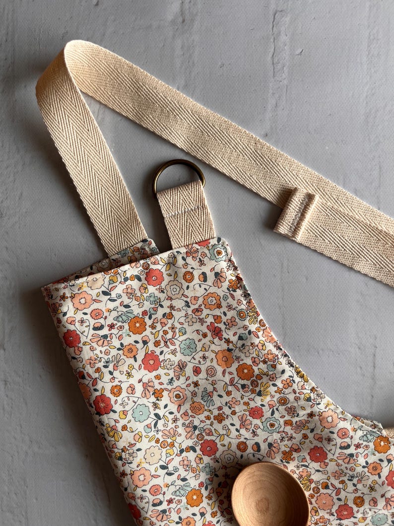 May include: An apron featuring a dense floral pattern with small red, orange, pink, and light blue flowers on a cream background. It includes wide, beige herringbone-patterned cotton straps and a bronze-colored metal D-ring for adjustment.