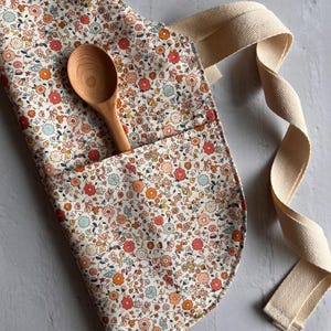 May include: A kitchen apron featuring a dense floral pattern in shades of orange, pink, and light blue on a cream background. The apron includes a front pocket containing a light-colored wooden spoon and features thick, cream-colored herringbone straps.