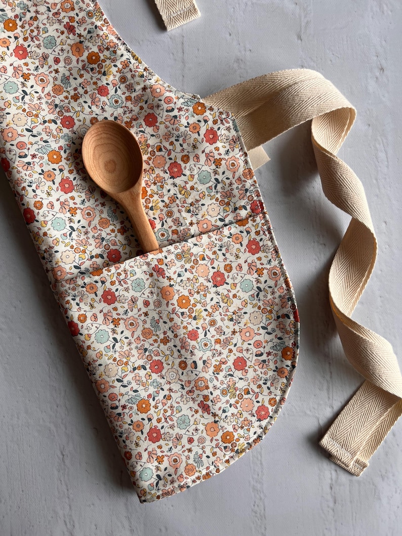 May include: A kitchen apron featuring a dense floral pattern in shades of orange, pink, and light blue on a cream background. The apron includes a front pocket containing a light-colored wooden spoon and features thick, cream-colored herringbone straps.
