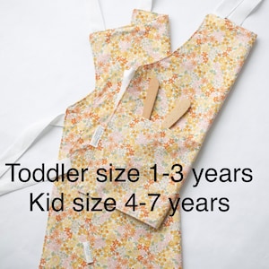 May include: Two floral print aprons with white straps. The smaller apron is labeled "Toddler size 1-3 years" and the larger apron is labeled "Kid size 4-7 years".