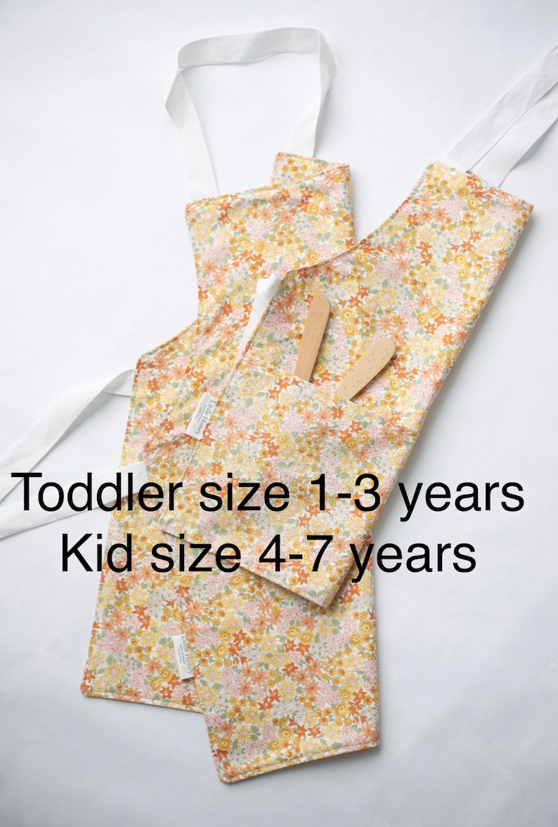 May include: Two floral print aprons with white straps. The smaller apron is labeled "Toddler size 1-3 years" and the larger apron is labeled "Kid size 4-7 years".
