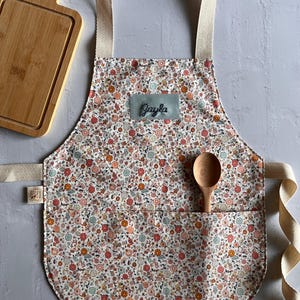 May include: A floral print apron with the name "Jayla" embroidered on a patch. The apron has a pocket with a wooden spoon inside. A wooden cutting board is in the background.