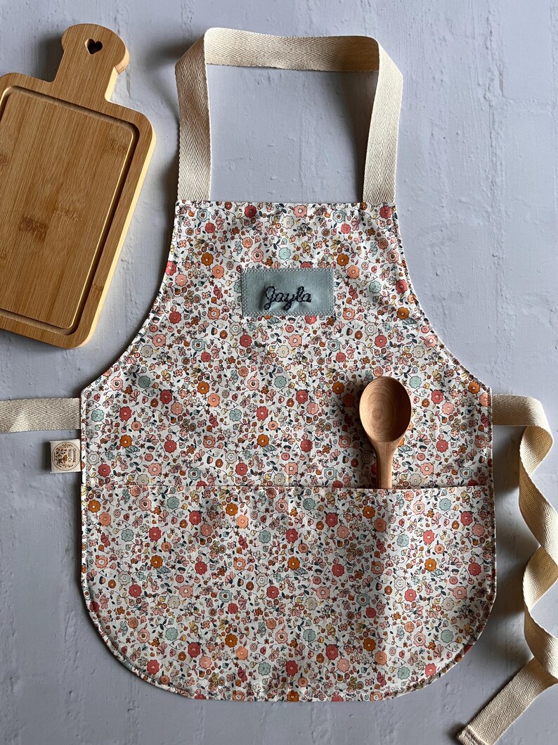 May include: A floral print apron with the name "Jayla" embroidered on a patch. The apron has a pocket with a wooden spoon inside. A wooden cutting board is in the background.