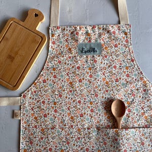 May include: A white apron with a floral print in shades of pink, orange, and blue. The apron has a large pocket on the front and a name tag that says "Estella".