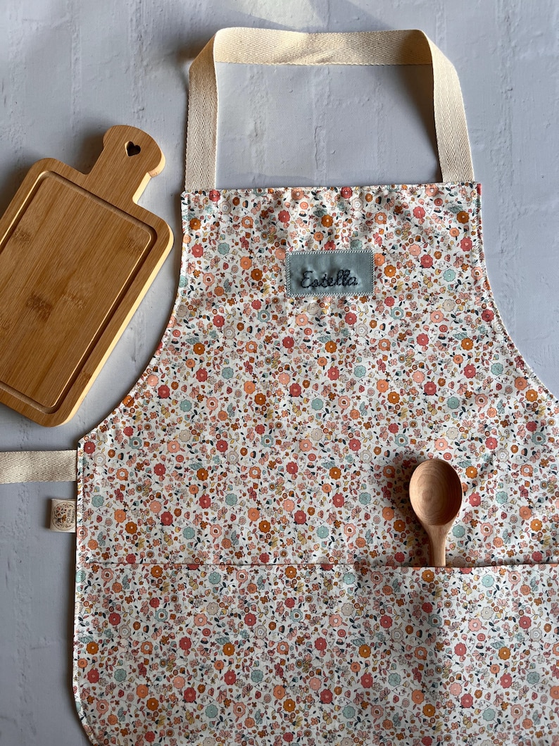 May include: A white apron with a floral print in shades of pink, orange, and blue. The apron has a large pocket on the front and a name tag that says "Estella".