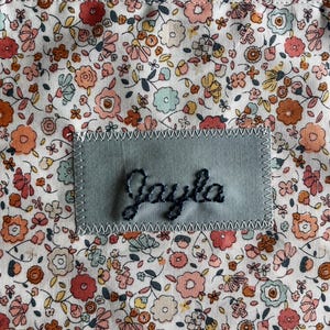 May include: A floral patterned fabric with a patch that reads "Jayla" in cursive. The fabric has a white background with small flowers in shades of pink, orange, yellow, and blue. The patch is a light blue-green color.