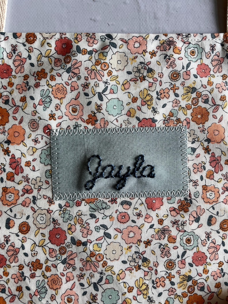 May include: A floral patterned fabric with a patch that reads "Jayla" in cursive. The fabric has a white background with small flowers in shades of pink, orange, yellow, and blue. The patch is a light blue-green color.