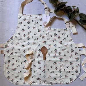 May include: Two cream-colored aprons featuring a repeating floral pattern of small pink, blue, and dark red flowers with green leaves. Each apron has a front pocket, adjustable beige neck straps, and a small wooden spoon tucked into the pocket.