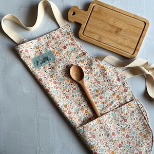 May include: A white apron with a floral print and a pocket. The apron has a cream-colored strap and the name "Estella" is embroidered on the front. A wooden spoon and a small wooden cutting board are also included.