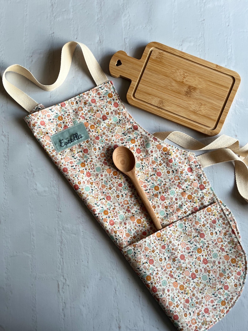 May include: A white apron with a floral print and a pocket. The apron has a cream-colored strap and the name "Estella" is embroidered on the front. A wooden spoon and a small wooden cutting board are also included.