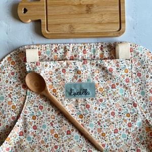 May include: A white apron with a floral print and a wooden spoon. The apron has a pocket and a name tag that says "Estella". There is also a wooden cutting board with a heart-shaped handle.