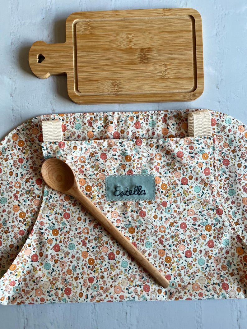 May include: A white apron with a floral print and a wooden spoon. The apron has a pocket and a name tag that says "Estella". There is also a wooden cutting board with a heart-shaped handle.
