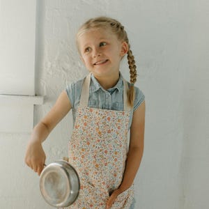 May include: A young person wearing a floral print apron and a blue denim shirt holds a silver cooking pot.