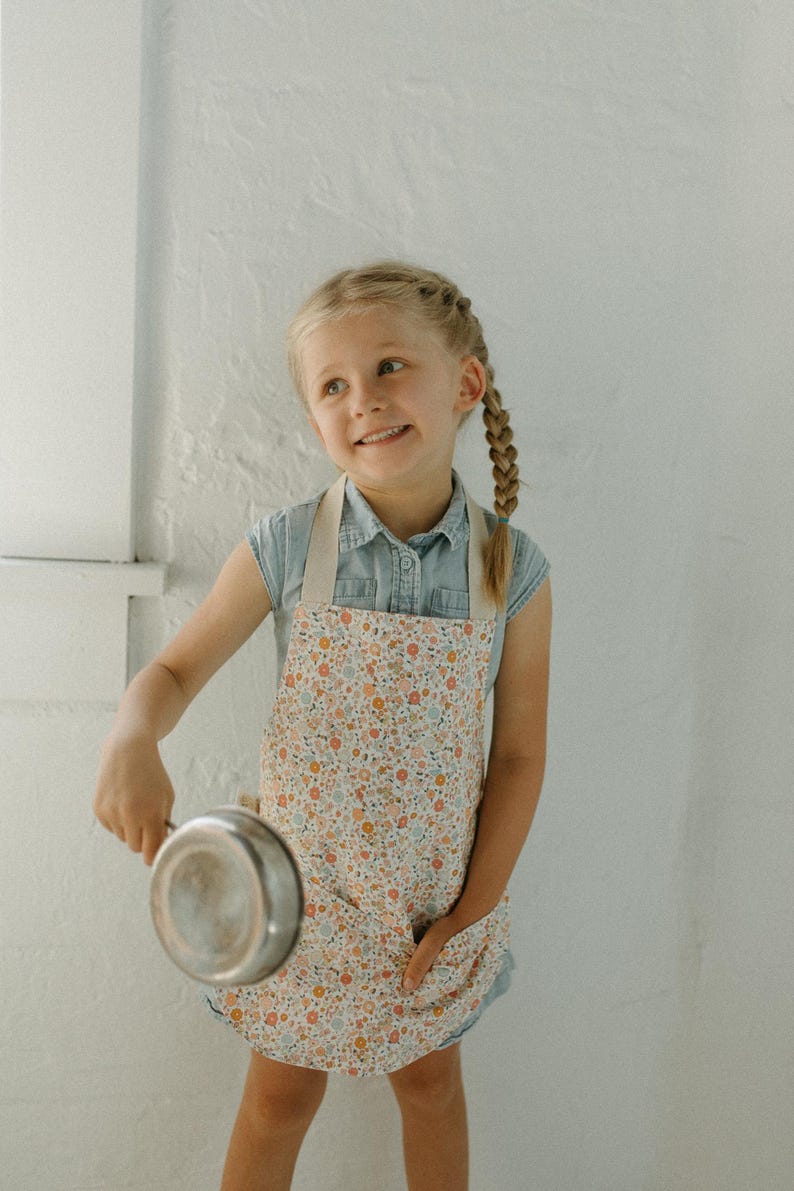 May include: A young person wearing a floral print apron and a blue denim shirt holds a silver cooking pot.