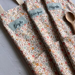 May include: Close-up of three floral print aprons with light blue name patches embroidered with the names "Jayla", "Ada Jane", and "Brynlee". Each apron has a pocket and a beige strap. A wooden spoon rests in one pocket.