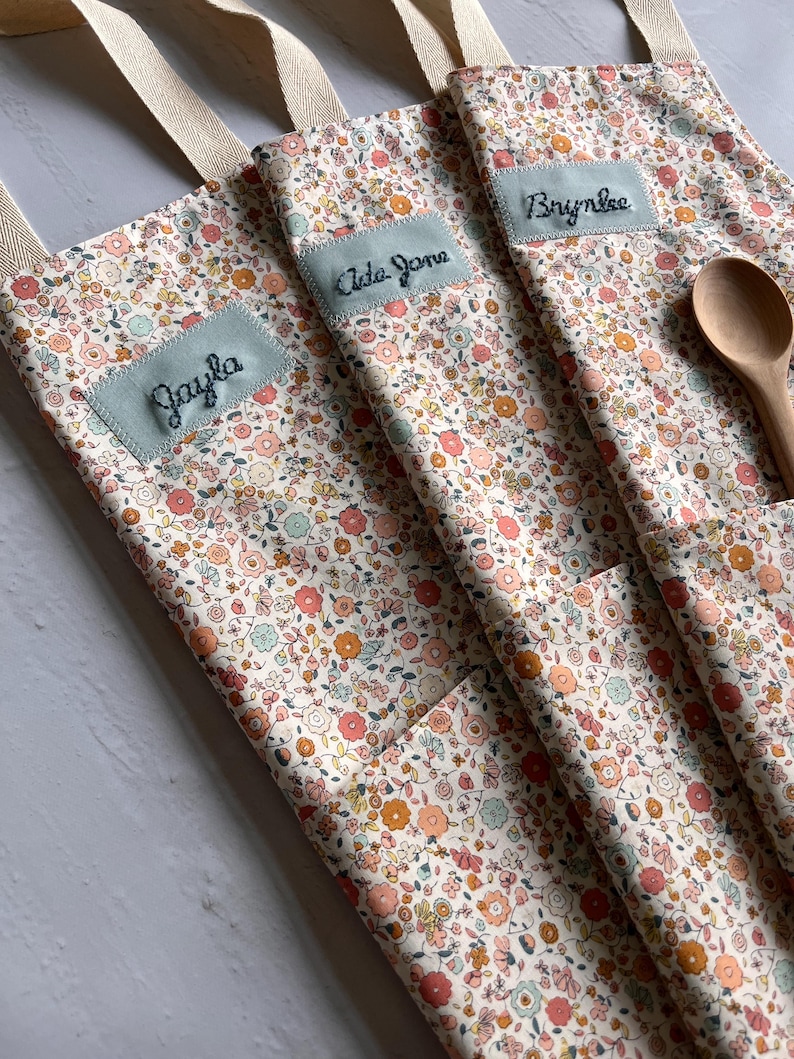 May include: Close-up of three floral print aprons with light blue name patches embroidered with the names "Jayla", "Ada Jane", and "Brynlee". Each apron has a pocket and a beige strap. A wooden spoon rests in one pocket.
