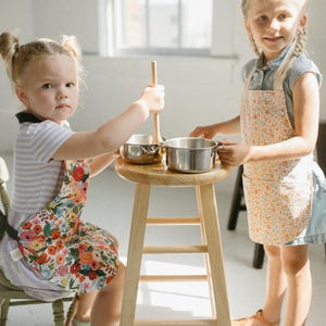 May include: Two children wearing floral print aprons are playing with toy pots and a wooden spoon in a kitchen setting. The child on the left is holding a wooden spoon and the child on the right is holding a pot.
