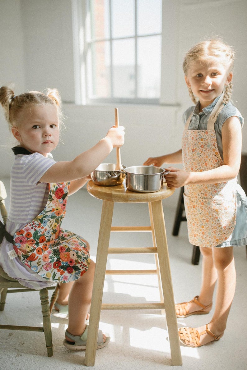 May include: Two children wearing floral print aprons are playing with toy pots and a wooden spoon in a kitchen setting. The child on the left is holding a wooden spoon and the child on the right is holding a pot.